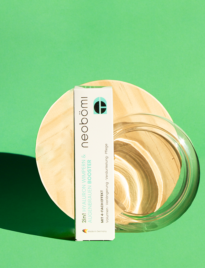 Produktverpackung neobömi 2in1 Wimpern & Augenbrauenbooster vor stehend vor einer runden Holzform.